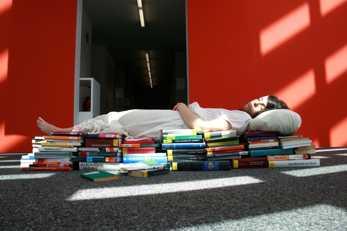 A woman with pale skin and dark brown hair is lying on many different stacks of books which make a bed for her. She is wearing a white gown and has a pillow under her head. The carpet on the ground is grey and shadows from the windows reflect on her body and on the carpet. Behind her are bright red walls and the entrance to a hallway that is lit up by flourescent lights. The woman's face looks peaceful and she seems to be sleeping serenely on her bed of books.