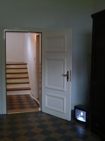 The image depicts a white hallway with checkered floor an. On the bottom right a corner there is a small television with a black and white image.