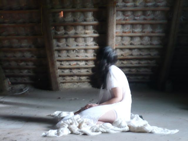 An blurry attic with the unfocussed figure of a woman with long dark hair sitting on white sheets.