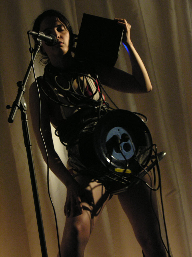 A woman with light skin and dark hair is holding a black box on her shoulder. A extension cord drum is wrapped around her waist and her body is wrapped with cables.