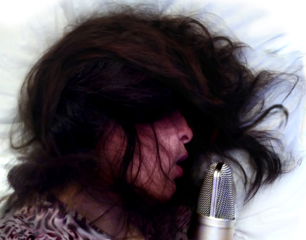 A profile of a female presenting person lying down with hair covering her eyes and face. There is a microphone in front of her mouth and she could be singing into it as she lies on her pillow.